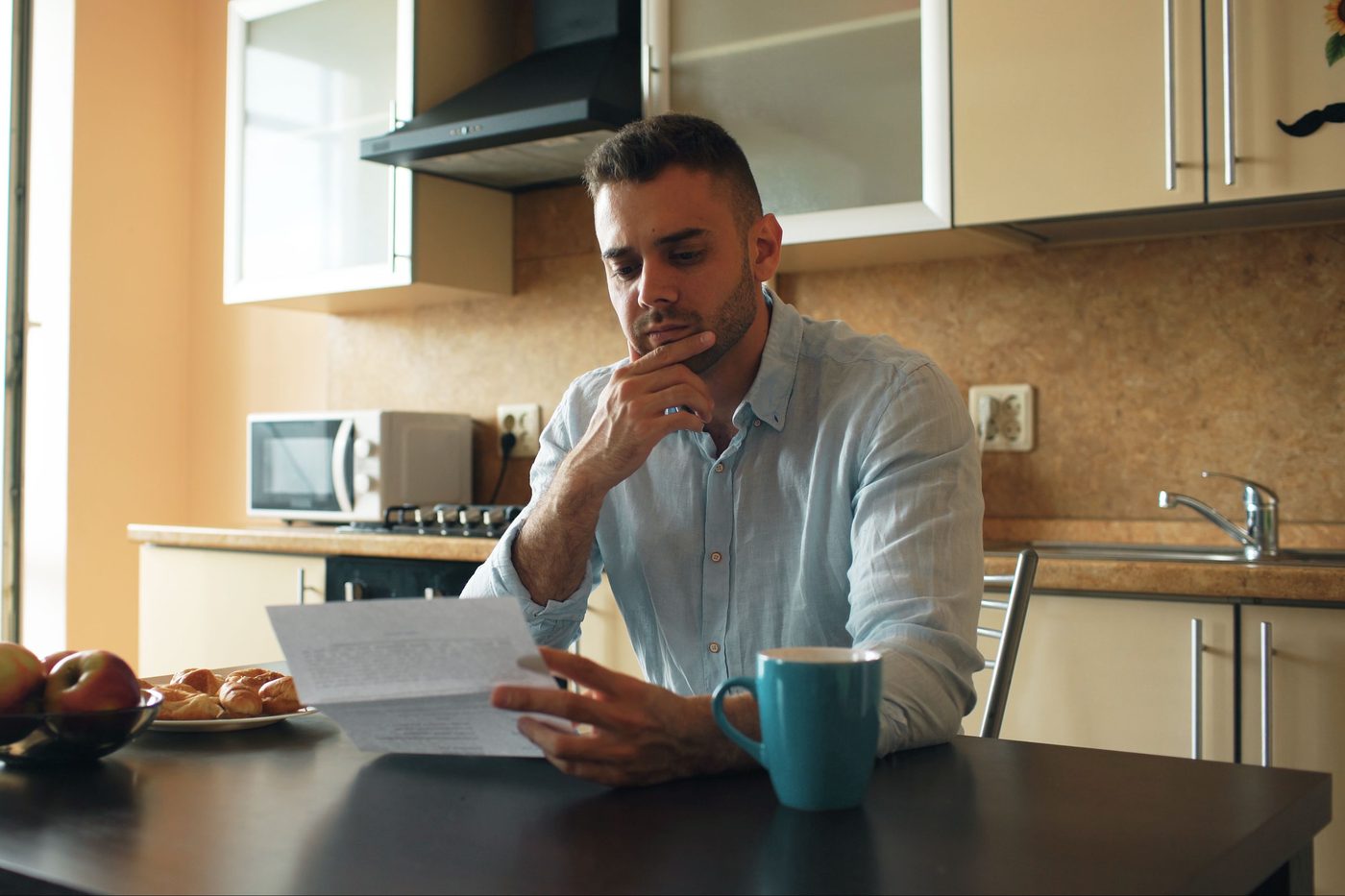 Man sitting at his kitchen table with a hand on his chin, reading a letter carefully